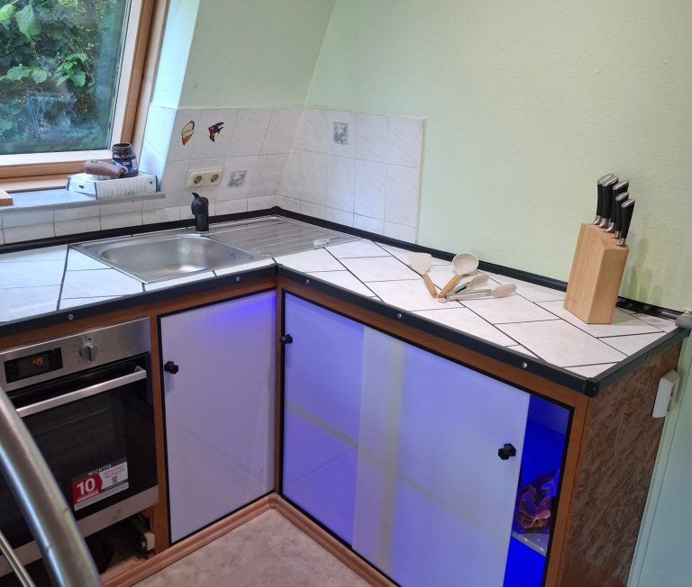Kueche2 Küche mit grauen Schränken, weißem Fliesenspiegel und blauer LED-Beleuchtung.