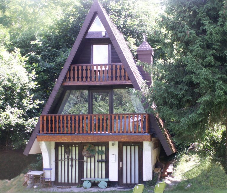 Bungalow neu Ein dreieckiges, zweigeschossiges Ferienhaus mit Balkon und umgebender grüner Vegetation.