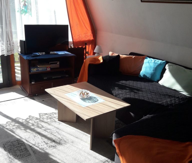 Stube Helle Wohnzimmereinrichtung mit Sofa, Tisch und Fernseher, orangefarbene Vorhänge.