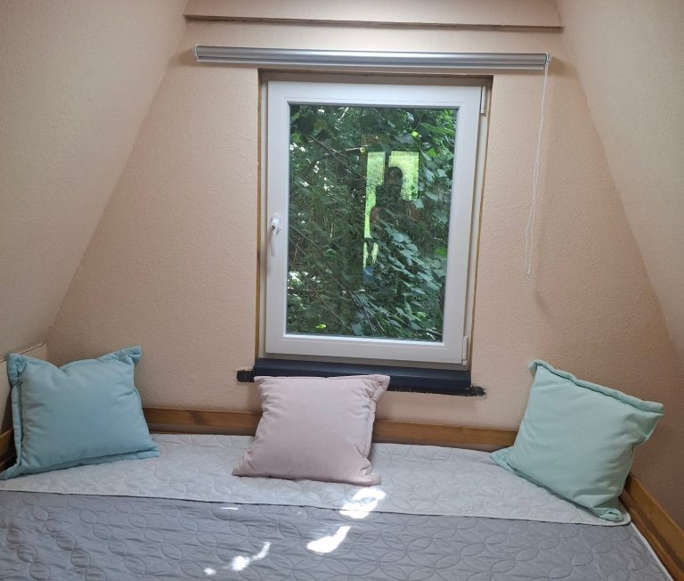 Schlafzimmer2 neu Fenster mit Blick auf Bäume, neben einem Doppelbett mit bunten Kissen in einem Dachzimmer.