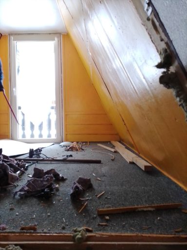 Unfertiger Raum mit beschädigter Wand, losem Bodenbelag und Trümmern auf dem Boden.