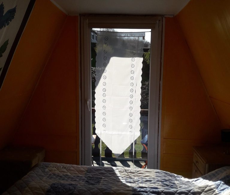 Schlfzimmer1 alt Blick aus einem Dachfenster auf eine helle Gardine und einem Bett.