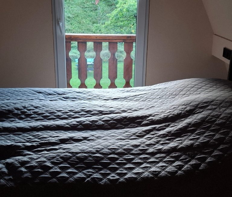 Gemütliches Schlafzimmer mit Bett, Fensterblick ins Grüne und einem Buch auf der Bettdecke.