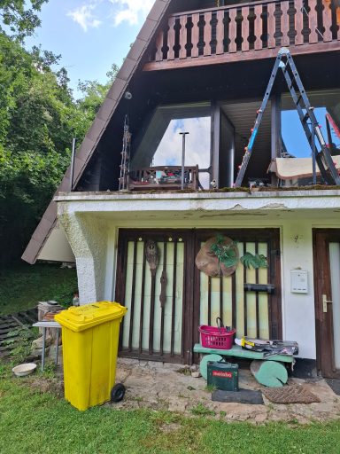 Haus mit Balkon, gelber Mülltonne und Werkzeug auf dem Boden, umgeben von Bäumen.